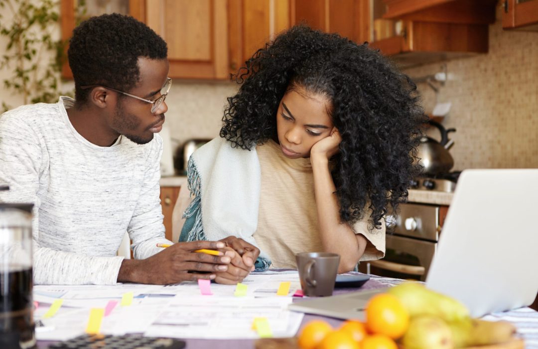 Young African family facing financial crisis. Husband in glasses trying to soothe his beautiful wife, holding her hand and telling that everything will be alright while managing finances in kitchen