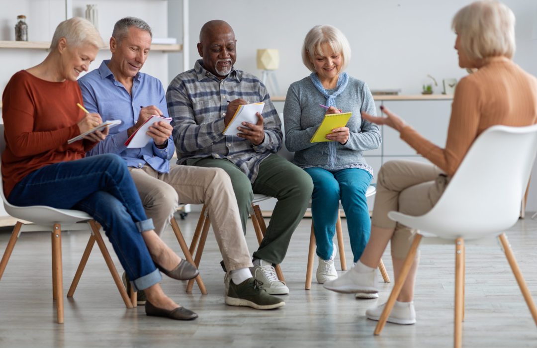 Multiracial group of cheerful elderly men and women in casual sitting on chairs in front of senior coach, taking notes, having educational class at retreat center or nursing home